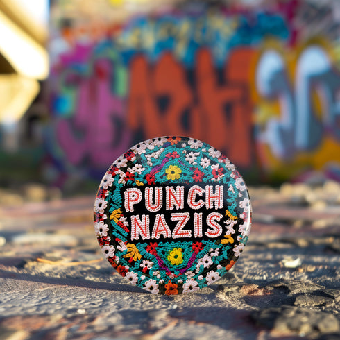 Round pinback metal button with an embroidered-style design on a black background. The center displays the phrase "PUNCH NAZIS" in bold, white stitched-textured lettering with red accents. Surrounding the text is an elaborate folk-inspired floral pattern, densely packed with embroidered-style flowers in shades of red, yellow, blue, pink, and white. The intricate design includes symmetrical lace-like stitching and decorative motifs, creating a rich, tapestry-like effect.